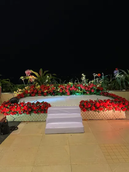 An outdoor stage setup for a red-themed event at night. The stage is beautifully decorated with a dense arrangement of red flowers, creating a bold and romantic focal point.