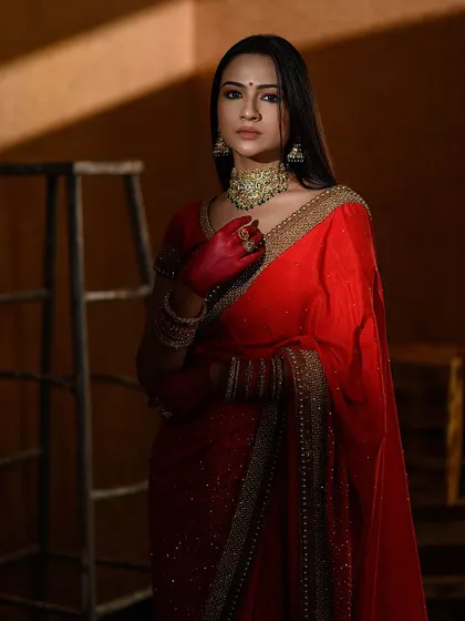 A dark and moody portrait in a red saree. The dramatic lighting from the side creates interesting shadows and highlights the model's features.