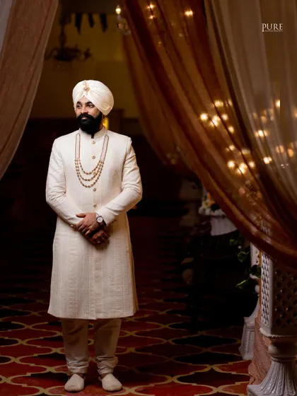 A striking portrait of the groom standing in a beautifully lit doorway. His elegant off-white sherwani and confident pose exude regal charm.