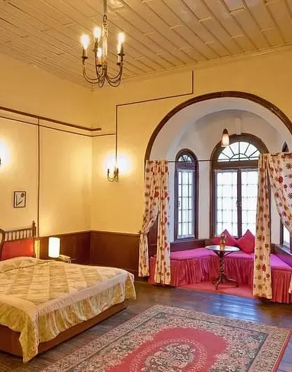 A spacious hotel suite featuring a classic bed, a cozy bay window seating area, and traditional furniture.