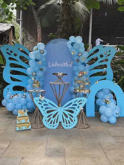 A stunning outdoor butterfly-themed party in shades of blue. The setup includes large laser-cut butterfly wing backdrops and elegant cake stands.