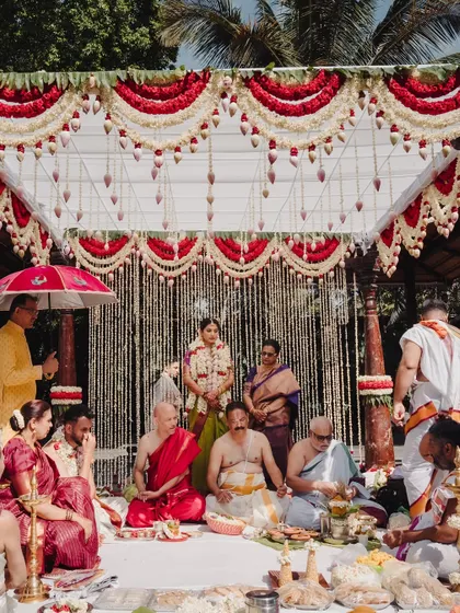 A traditional South Indian wedding ceremony in progress, set against a backdrop of red and white floral decorations. We manage every detail of the rituals to ensure a seamless and meaningful experience for the family.