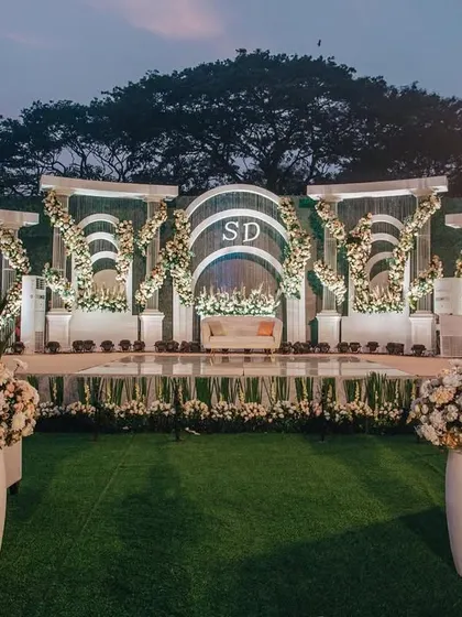 A grand reception stage with neoclassical arches and columns, adorned with cascading white floral arrangements. The monogram in the center adds a personal touch to the majestic design.