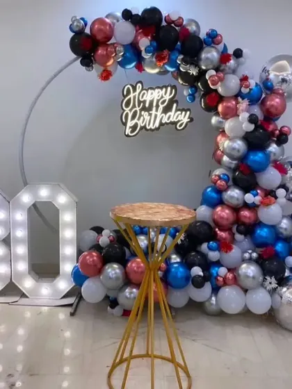 A vibrant 30th birthday party decoration. A circular frame is adorned with a colorful mix of red, blue, black, and silver balloons, complemented by a neon sign and light-up '30' numbers.