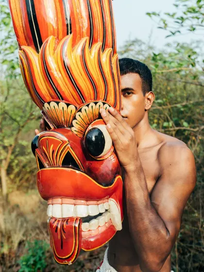 A portrait from the "Nazaar" series, where the model's face is partially obscured by a large, colorful traditional mask. The outdoor setting adds an earthy, raw quality.