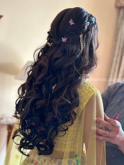 A whimsical half-updo with soft curls, adorned with delicate butterfly clips and a sparkling gold accessory. This is a perfect hairstyle for a dreamy and romantic Mehendi look.
