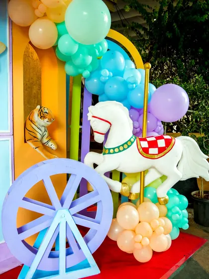 A classic carousel horse and other carnival props set against a rainbow backdrop with pastel balloons.