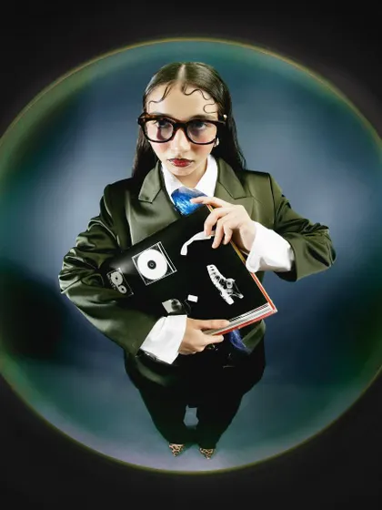 A fisheye portrait of a model styled in a green suit and glasses, holding a book. The distorted, high-angle perspective creates a quirky, intellectual character within a surreal visual world.