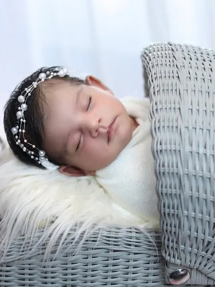 A close-up of a sleeping baby in a vintage-style grey wicker pram. The pearl headband adds a touch of classic elegance to this simple and sweet portrait.