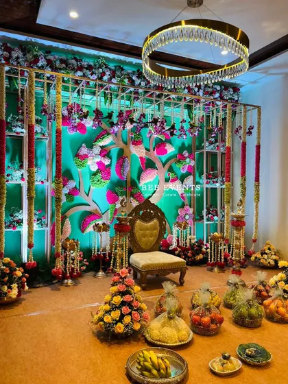 A beautiful and intricate stage setup for a traditional ceremony. The 'tree of life' backdrop, adorned with colorful flowers and surrounded by fruit baskets, creates a vibrant and auspicious setting.