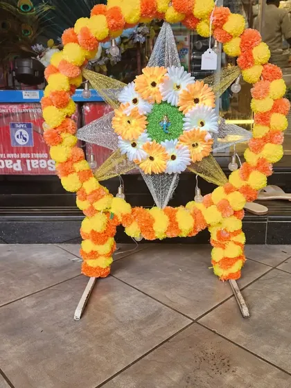 A beautiful circular Ganapati backdrop frame decorated with artificial marigold garlands in yellow and orange. The center features a star-shaped design with gerbera daisies, creating a traditional and auspicious look.