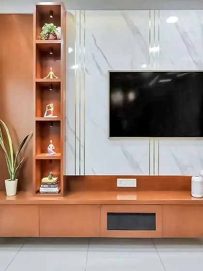 A floating TV unit in a warm wood finish set against a white marble-patterned wall with gold inlay details. The design includes a vertical display cabinet with internal lighting, adding a touch of elegance.