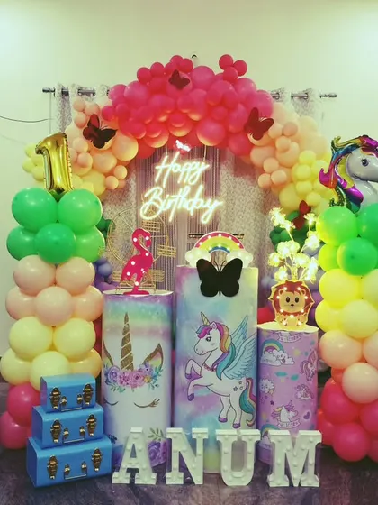 A complete unicorn party stage with a rainbow balloon arch and themed plinths. The setup is brightened by a neon sign and includes marquee letters spelling out the birthday girl's name, creating a perfect fantasy setting.