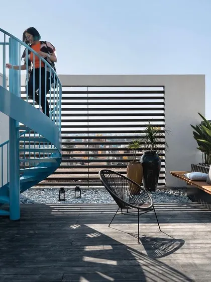 The upper terrace features a bright blue spiral staircase that acts as a sculptural element. The design includes a wooden deck, built-in seating with planters, and modern outdoor furniture, creating a perfect space for relaxing in the sun.