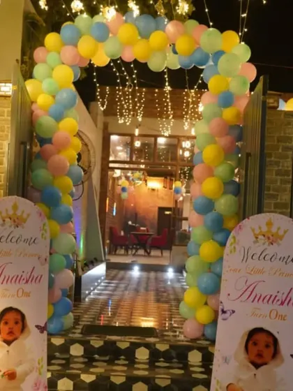 The entrance to a first birthday party, decorated with a pastel balloon arch and personalized welcome stands featuring the baby's photo.
