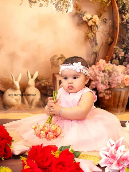 A lovely portrait from a 6-month-old's sitter session. Dressed in a soft pink tulle dress, she is sitting in a whimsical studio setting decorated with flowers and adorable bunny figurines, creating a gentle, storybook feel.