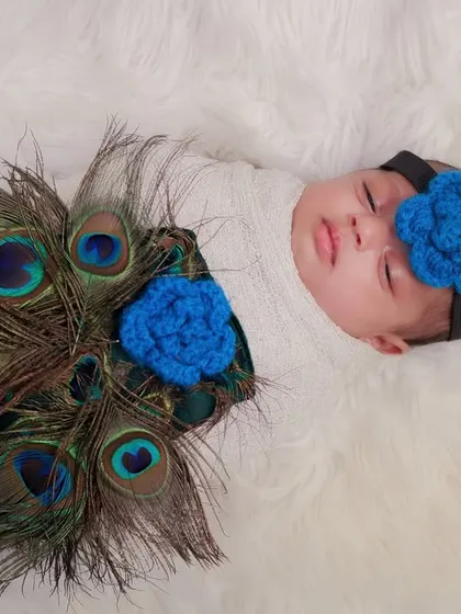 A beautiful overhead shot of a newborn decorated with peacock feathers, creating a stunning visual pattern. The blue knitted flowers add a lovely touch of color.