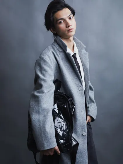 A stylish studio portrait for a male model. The combination of the grey overcoat and geometric backpack creates a modern, high-fashion look.