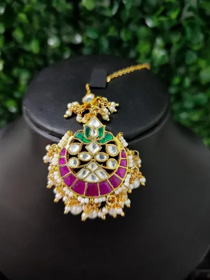 A unique maang tikka with a crescent design in magenta enamel and kundan work. The pearl drops add a soft touch.