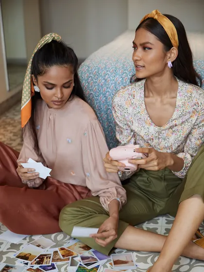 A lifestyle shot that tells a story of friendship and nostalgia. The models are looking at polaroids, creating a warm, relatable scene that showcases the clothing in a relaxed, everyday context.