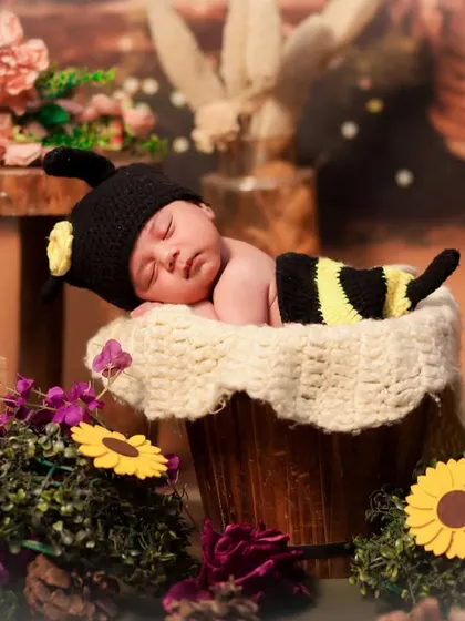 A newborn in a bee costume sleeps in a bucket filled with soft fabric, surrounded by sunflowers and other floral elements for a garden feel.