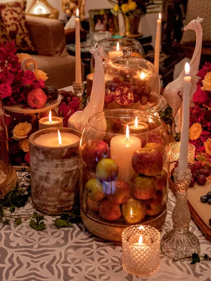 A rich and moody tablescape perfect for a wine and cheese evening. The decor features an abundance of candles, fresh fruits like apples and grapes, and deep red florals, creating a warm, rustic, and opulent still life.