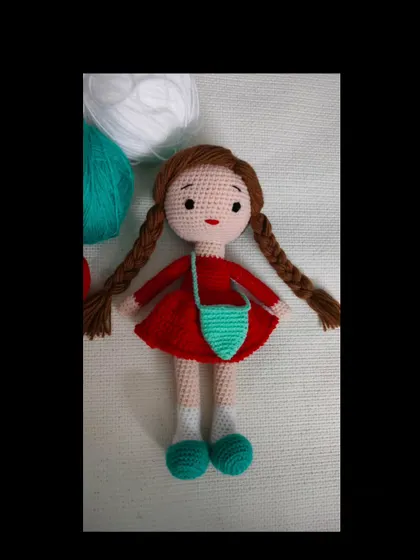 A handmade crochet doll with two long braids, a red dress, and a little green bag.