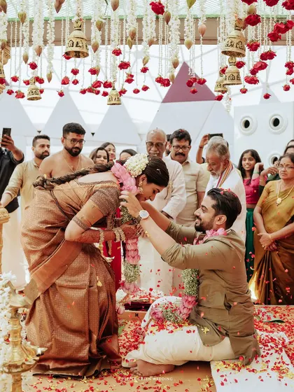The groom, Vinay, places the garland on his bride, Sradha, in a moment of sacred union. The scene is full of tradition, emotion, and the blessings of family.