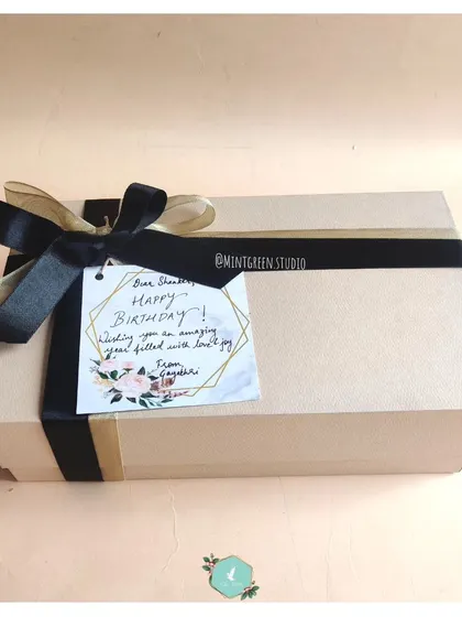 A beautifully wrapped birthday gift box. The simple cardboard box is elevated with elegant black and gold ribbons and a custom handwritten tag, showing that even simple packaging can be made special.