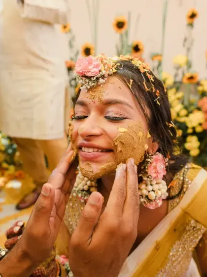 A close-up of the bride's smiling face as she gets haldi applied. The vibrant yellow paste and flower jewellery create a beautiful, traditional Haldi portrait.