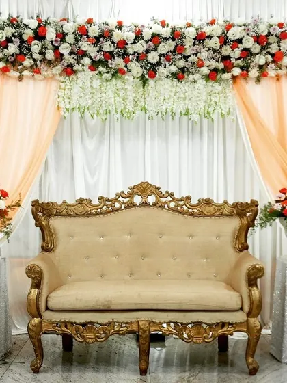 This engagement stage decoration features a classic sofa set against a backdrop of white and peach drapes. The top is adorned with a thick arrangement of red and white flowers, creating a simple yet elegant look.