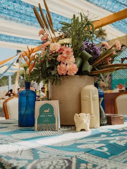 A close-up of a tablescape, featuring a hand-blocked print, blue glass bottles, and a rustic floral arrangement. The design feels both elegant and grounded in local craft.