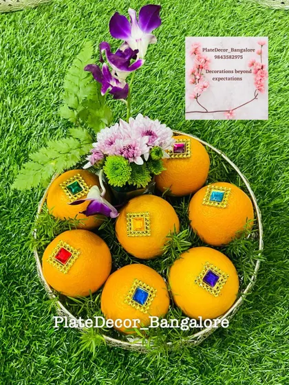 Oranges decorated with square kundan stones in multiple colors, arranged with fresh flowers in a silver bowl.