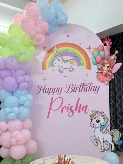 A beautiful unicorn-themed birthday backdrop for Prisha. The setup features a pastel rainbow balloon arch over a custom printed backdrop with unicorns and fairies.