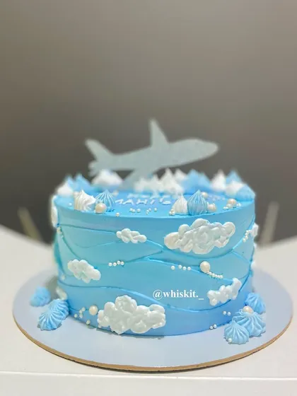 A sky-themed cake with fluffy white clouds and an airplane topper. The wavy blue frosting gives the impression of flying through the air.