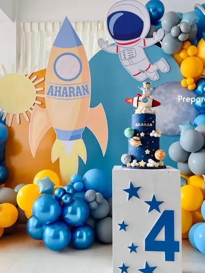 A fantastic outer space theme for a fourth birthday. The setup includes a rocket ship, an astronaut cutout, planet-themed balloons, and a custom cake plinth with the birthday boy's name.