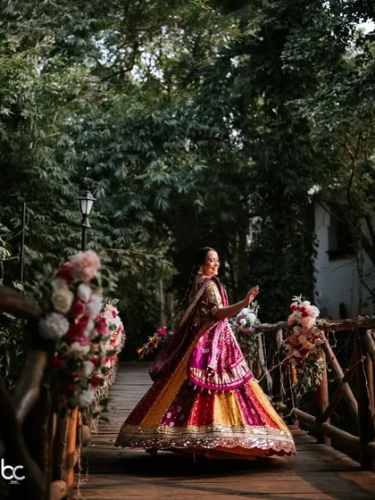 A beautiful shot of the bride twirling in her vibrant, multi-colored lehenga on a wooden bridge surrounded by lush greenery.