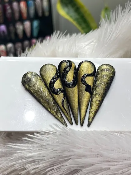 This set of five stiletto nail tips displays an impressive hand-painted black snake design over a textured gold and black gradient background. It's a powerful and luxurious example of our custom, high-fashion nail art.