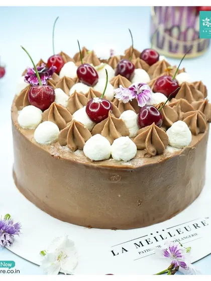 A classic Black Forest cake, generously decorated with fresh, juicy cherries, chocolate cream swirls, and dollops of white cream. The sides are coated in smooth chocolate frosting for a rich and satisfying experience.