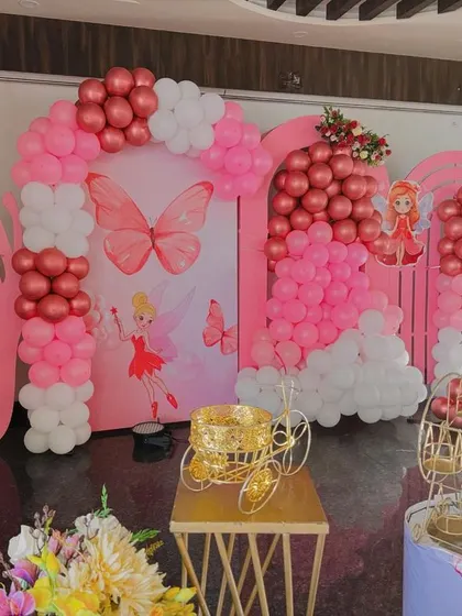 A charming pink butterfly-themed setup perfect for a first birthday. The decor includes large butterfly wing cutouts, balloon arches, and fairy-themed props.