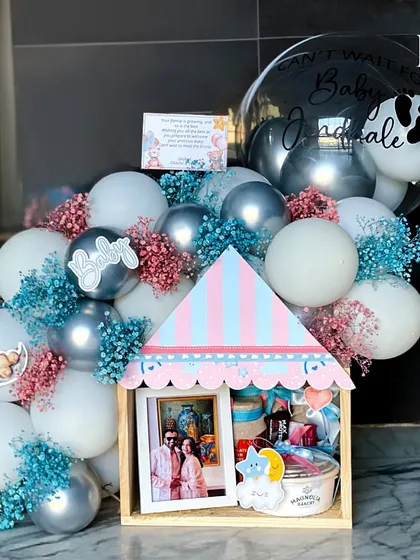 A hamper designed to celebrate the parents-to-be. This house-shaped box is filled with treats and a photo of the happy couple, surrounded by a festive arrangement of pink and blue balloons.