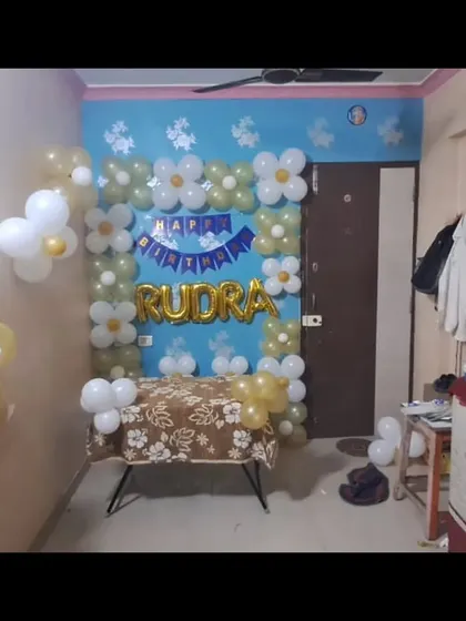 A simple birthday decoration at home with a 'Happy Birthday Rudra' foil balloon and a frame of white and gold balloons.
