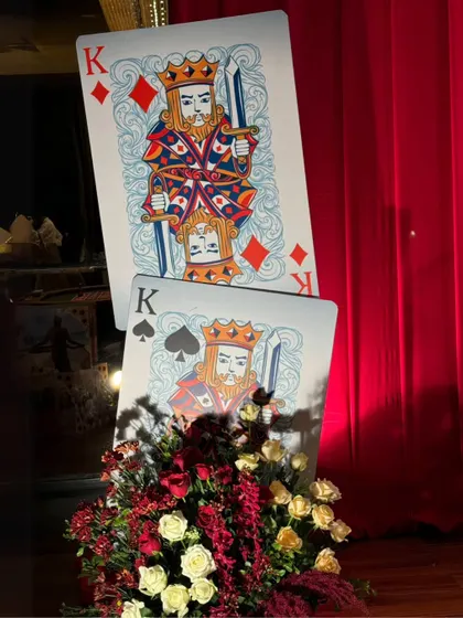 A detailed shot of the oversized playing card props, flanked by lush floral arrangements of red and white roses, adding a touch of softness to the bold theme.