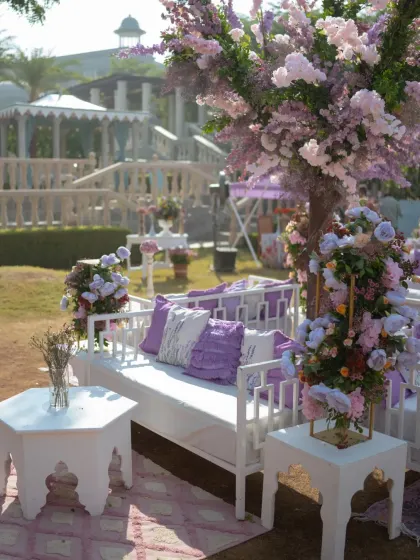 A serene seating area under a lavender-blossom tree, with white benches and plush purple cushions.