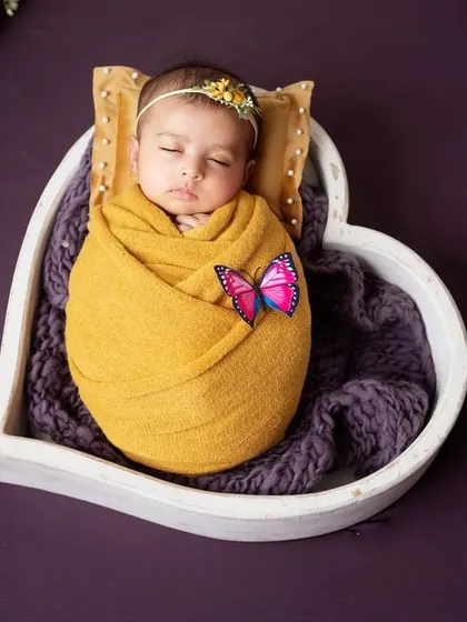 A newborn girl wrapped in a mustard yellow swaddle sleeps in a heart-shaped bowl, decorated with small butterflies.
