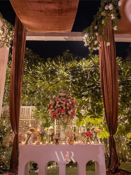 A welcome console table decorated with a large floral arrangement and surrounded by candle stands. The rich brown drapes and hanging chandeliers frame the space beautifully.