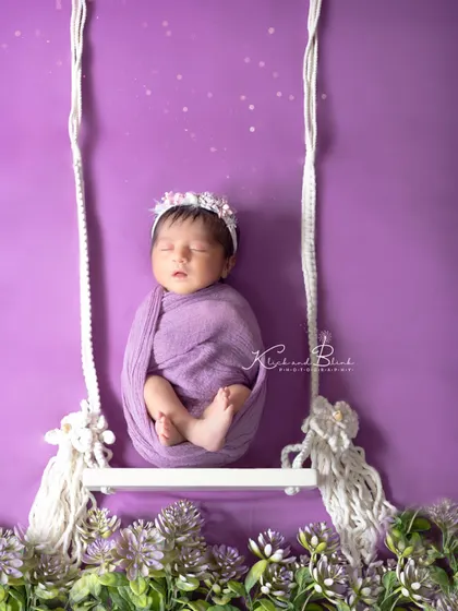 Swing me to the moon. A baby wrapped in lavender rests on a tiny swing against a purple backdrop, creating a dreamy and imaginative portrait.