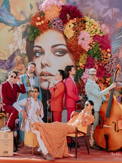 An editorial-style group portrait of the couple with their vibrant band against a colorful, artistic backdrop during their carnival event at Umaid Bhawan.