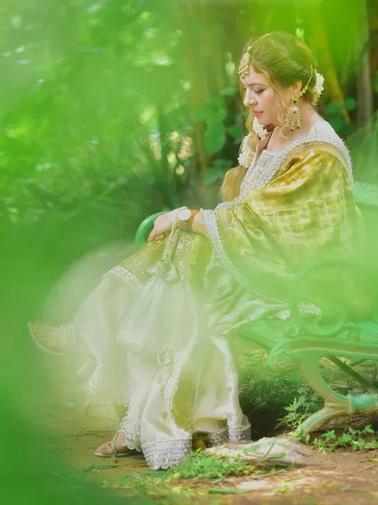 A creative shot using foreground blur to create a soft, dreamy effect. The focus is on her serene expression as she sits on a bench, surrounded by nature.