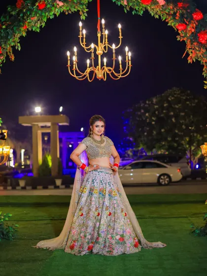 A full-length portrait of the bride under a grand floral arch and chandelier. This shot showcases her entire stunning outfit and the beautiful outdoor evening decor.